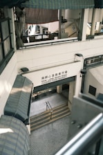 A multi-level market building with a sign reading 'Nam Long Shan Road Cooked Food Market'. The architecture features covered awnings and staircases leading downwards. The interior appears utilitarian with metal railings and tiled flooring.