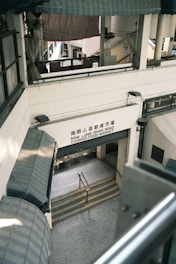 A multi-level market building with a sign reading 'Nam Long Shan Road Cooked Food Market'. The architecture features covered awnings and staircases leading downwards. The interior appears utilitarian with metal railings and tiled flooring.