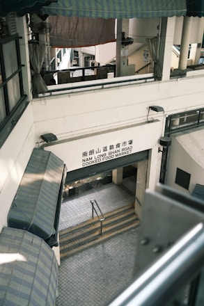 A multi-level market building with a sign reading 'Nam Long Shan Road Cooked Food Market'. The architecture features covered awnings and staircases leading downwards. The interior appears utilitarian with metal railings and tiled flooring.