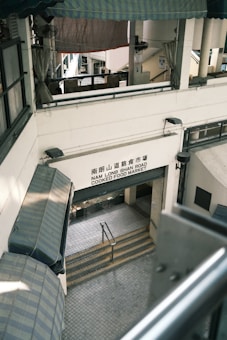 A multi-level market building with a sign reading 'Nam Long Shan Road Cooked Food Market'. The architecture features covered awnings and staircases leading downwards. The interior appears utilitarian with metal railings and tiled flooring.