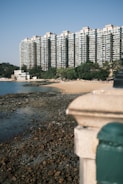 A large residential building complex with multiple stories towers over a small, rocky beach area lined with greenery. The clear sky and the water lapping onto the rocky shoreline create a serene and tranquil atmosphere.