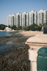 A large residential building complex with multiple stories towers over a small, rocky beach area lined with greenery. The clear sky and the water lapping onto the rocky shoreline create a serene and tranquil atmosphere.