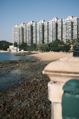 A large residential building complex with multiple stories towers over a small, rocky beach area lined with greenery. The clear sky and the water lapping onto the rocky shoreline create a serene and tranquil atmosphere.
