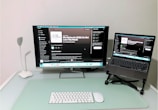 A modern and minimalistic home office setup with a desk featuring a large monitor displaying a podcast on a streaming platform, accompanied by a laptop on a stand showing the same content. A sleek white desk lamp is positioned on the left, with a wireless keyboard and mouse placed on a mint-colored desk pad.