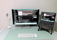 A modern and minimalistic home office setup with a desk featuring a large monitor displaying a podcast on a streaming platform, accompanied by a laptop on a stand showing the same content. A sleek white desk lamp is positioned on the left, with a wireless keyboard and mouse placed on a mint-colored desk pad.