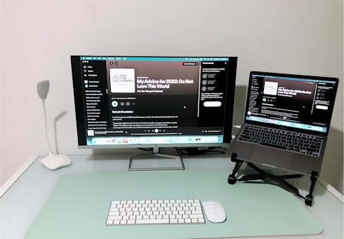 A modern and minimalistic home office setup with a desk featuring a large monitor displaying a podcast on a streaming platform, accompanied by a laptop on a stand showing the same content. A sleek white desk lamp is positioned on the left, with a wireless keyboard and mouse placed on a mint-colored desk pad.