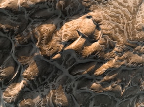 An aerial view of a rugged landscape showcasing intricately carved earth patterns and elevation changes, with light and shadow creating dramatic contrasts. The surface appears weathered with textures that resemble both rock formations and sandy dunes.