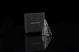 A black box with the brand name 'COPPERIST.WU' is positioned next to a metallic, sculptural object. The object resembles a stack of polished, reflective stones or a stylized scale pattern, and both items are set against a dark, glossy surface, creating a mirrored reflection beneath them.