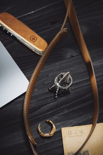 A close-up view of a wooden-handled multitool, brown leather belt, a metallic ring, a detailed metallic pendant, and an old piece of paper with 'USA' written on it, all placed on a dark wooden surface.