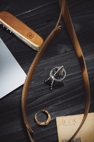 A close-up view of a wooden-handled multitool, brown leather belt, a metallic ring, a detailed metallic pendant, and an old piece of paper with 'USA' written on it, all placed on a dark wooden surface.
