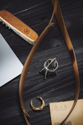 A close-up view of a wooden-handled multitool, brown leather belt, a metallic ring, a detailed metallic pendant, and an old piece of paper with 'USA' written on it, all placed on a dark wooden surface.