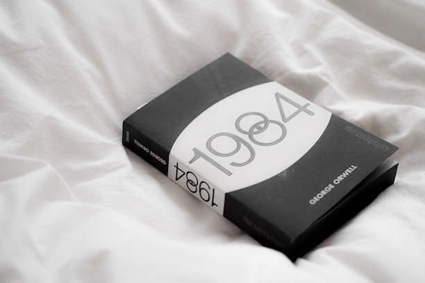 A black paperback book titled '1984' by George Orwell rests on crumpled white sheets. The cover design features a minimalistic style with monochrome tones.