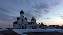 An Orthodox church with distinctive onion domes stands amidst a snowy landscape. The sky is filled with scattered clouds and a subtle gradient of colors from the setting or rising sun.