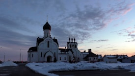An Orthodox church with distinctive onion domes stands amidst a snowy landscape. The sky is filled with scattered clouds and a subtle gradient of colors from the setting or rising sun.