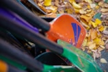Several colorful canoe paddles are piled closely together, surrounded by a scattering of yellow and brown autumn leaves on the ground.