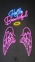 Neon sign art consisting of the words 'Hello Beautiful' in bright blue and pink letters. An illuminated yellow halo is positioned above two vibrant pink angel wings, creating a striking visual impact on a dark background.
