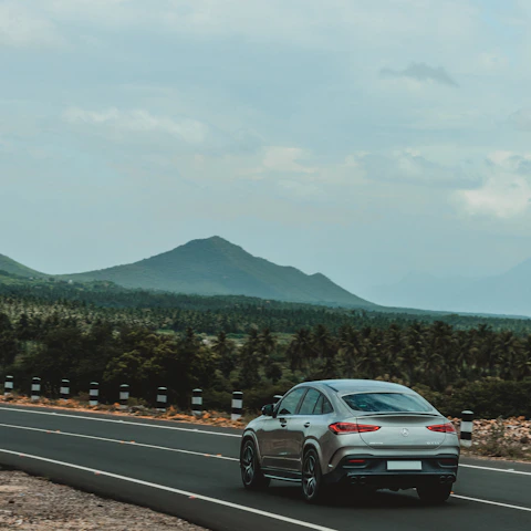 A sleek silver sedan cruising on a scenic Indian highway with lush greenery and hills in the background.