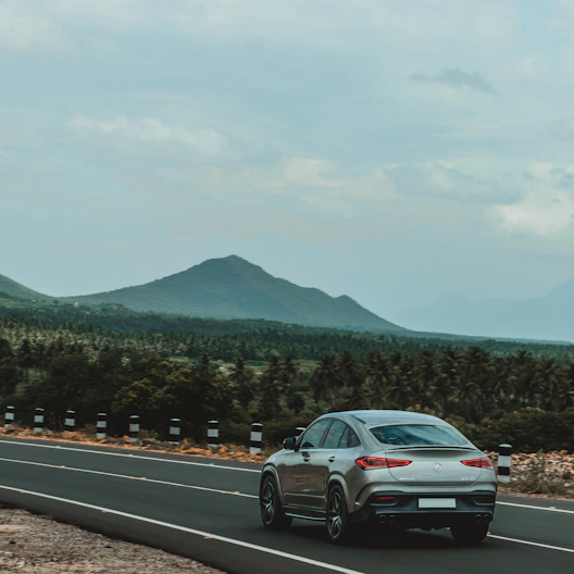 A sleek silver sedan cruising on a scenic Indian highway with lush greenery and hills in the background.