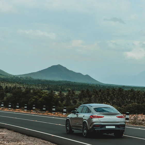A sleek silver sedan cruising on a scenic Indian highway with lush greenery and hills in the background.