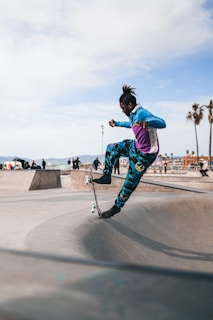 A person is skateboarding in mid-air at a skatepark. They are wearing colorful, athletic clothing, including a blue and purple jacket and patterned pants. The skatepark features various ramps and obstacles, and the background shows a clear sky with a few wispy clouds and palm trees silhouetted against the horizon.