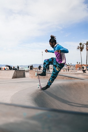 A person is skateboarding in mid-air at a skatepark. They are wearing colorful, athletic clothing, including a blue and purple jacket and patterned pants. The skatepark features various ramps and obstacles, and the background shows a clear sky with a few wispy clouds and palm trees silhouetted against the horizon.