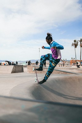 A person is skateboarding in mid-air at a skatepark. They are wearing colorful, athletic clothing, including a blue and purple jacket and patterned pants. The skatepark features various ramps and obstacles, and the background shows a clear sky with a few wispy clouds and palm trees silhouetted against the horizon.