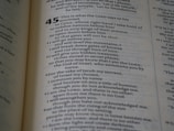 A close-up view of a book page showing text. The page contains a passage from a religious scripture, with verse numbers and text aligned in a structured format. The text is clearly printed in black ink on a light-colored background.