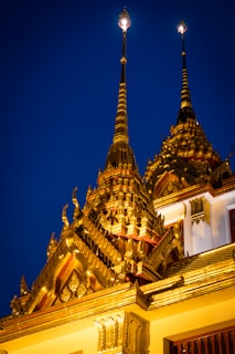 Elaborate gold spires of a Thai temple set against a deep blue evening sky. The architecture showcases intricate details with ornate patterns and sharp, pointed tops that reflect a bright, golden hue.