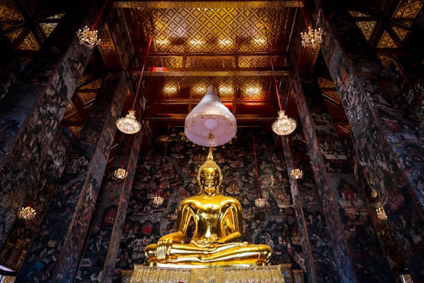 A grand and ornate interior scene featuring a large golden Buddha statue sitting in a traditional meditation pose, surrounded by intricately decorated walls and a richly adorned ceiling. Multiple hanging chandeliers illuminate the space, highlighting the elaborate patterns and artwork on the walls and ceiling. The atmosphere evokes a sense of reverence and peace.