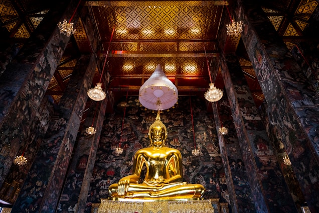 A grand and ornate interior scene featuring a large golden Buddha statue sitting in a traditional meditation pose, surrounded by intricately decorated walls and a richly adorned ceiling. Multiple hanging chandeliers illuminate the space, highlighting the elaborate patterns and artwork on the walls and ceiling. The atmosphere evokes a sense of reverence and peace.