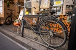 A vintage bicycle with a worn saddle is parked in front of a shop window. The window features decals of a giraffe and the words 'Cafe Giraffe.' There is a small orange chair and a sign with menu items displayed beside the bicycle. Another bicycle is visible inside the cafe behind the glass. The scene is lively with the reflection of street activity in the glass.
