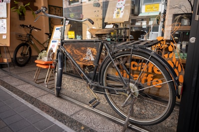 A vintage bicycle with a worn saddle is parked in front of a shop window. The window features decals of a giraffe and the words 'Cafe Giraffe.' There is a small orange chair and a sign with menu items displayed beside the bicycle. Another bicycle is visible inside the cafe behind the glass. The scene is lively with the reflection of street activity in the glass.