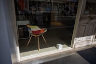 An indoor scene features a colorful patchwork chair with wooden legs positioned near a large window. Outside, a person is seen cycling, partially obscured by reflections and the glass. The space has a rustic, vintage feel with worn wooden doors and a muted concrete floor. Some small white box-like object sits on the ground nearby.