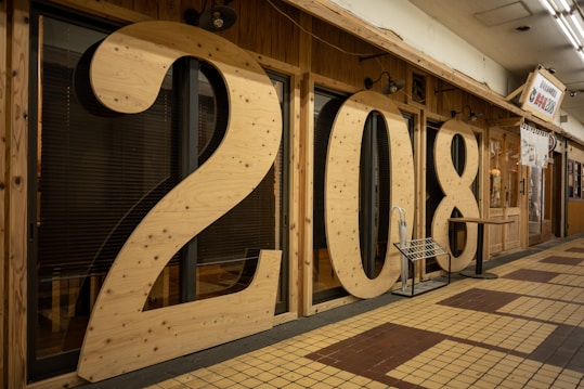 Large wooden numbers '208' are displayed prominently as part of the exterior decor of a shop or building. The numbers are mounted against a wooden wall, which features a pattern of dark horizontal blinds. The floor is covered with tiles in shades of yellow and brown, adding contrast to the scene.