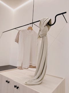 A minimalist clothing display with a white t-shirt on a wooden hanger and a beige knit scarf draped over a sleek black metal rod. The background is a clean, light-colored wall with subtle architectural lines.