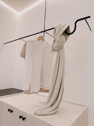 A minimalist clothing display with a white t-shirt on a wooden hanger and a beige knit scarf draped over a sleek black metal rod. The background is a clean, light-colored wall with subtle architectural lines.