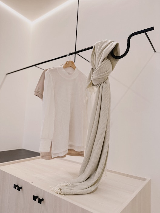 A minimalist clothing display with a white t-shirt on a wooden hanger and a beige knit scarf draped over a sleek black metal rod. The background is a clean, light-colored wall with subtle architectural lines.