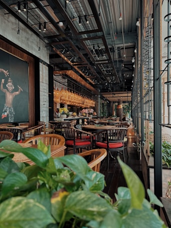 The interior of a cozy restaurant features wooden tables and chairs with red cushions. There are lush green plants in the foreground. The ceiling showcases industrial elements with exposed beams and lighting. Warm lighting from hanging lamps creates an inviting ambiance. A large painting hangs on the wall, adding an artistic touch.