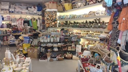 A colorful souvenir shop filled with various items including textiles, ceramics, clothing, hats, and bags. The shelves are densely packed with products like postcards, magnets, and other decorative pieces. Bright lighting highlights the vibrant assortment of items.