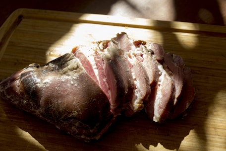 Sliced cured meat rests on a wooden cutting board, illuminated by natural light, casting shadows and highlighting the texture and marbling of the meat.