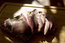 Sliced cured meat rests on a wooden cutting board, illuminated by natural light, casting shadows and highlighting the texture and marbling of the meat.