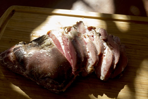 Sliced cured meat rests on a wooden cutting board, illuminated by natural light, casting shadows and highlighting the texture and marbling of the meat.