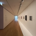 A modern art gallery hallway with wooden flooring and white walls featuring various abstract paintings. The ceiling has a row of lights illuminating the artwork. The corridor stretches towards a large window at the end.