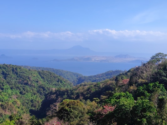A vibrant landscape displaying lush green hills and a serene blue lake set against the backdrop of distant mountains under a bright blue sky. The area is rich with diverse vegetation, demonstrating a mix of dense forest and open spaces.