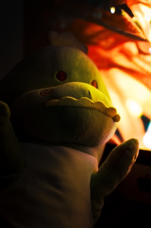 A soft, plush toy resembling a green dinosaur is sitting in front of a warm, glowing light source. The light casts a yellow hue on the toy, accentuating its round eyes and stitched-on details. The background is predominantly dark, creating a contrast with the illuminated areas.
