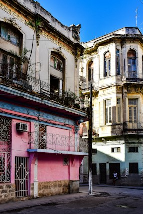 Colorful, weathered buildings with intricate ironwork and architectural details line a street corner, reflecting a mix of faded pastels and vibrant shades under a clear blue sky.