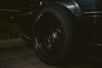A close-up view of a car tire mounted on a black alloy wheel featuring the BMW logo. The vehicle is parked near a curb with some greenery visible in the background. The image is taken in low-light conditions, emphasizing the sleek, polished appearance of the wheel and the tire tread.
