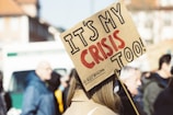 A crowd of people appears to be gathered, with a prominent sign in the foreground reading 'IT'S MY CRISIS TOO!' in bold letters. The sign is held by someone with long hair, and the text on the sign conveys a message of personal involvement or activism. The background shows more people slightly out of focus, indicating a group setting or protest.