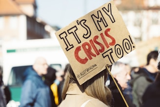 A crowd of people appears to be gathered, with a prominent sign in the foreground reading 'IT'S MY CRISIS TOO!' in bold letters. The sign is held by someone with long hair, and the text on the sign conveys a message of personal involvement or activism. The background shows more people slightly out of focus, indicating a group setting or protest.