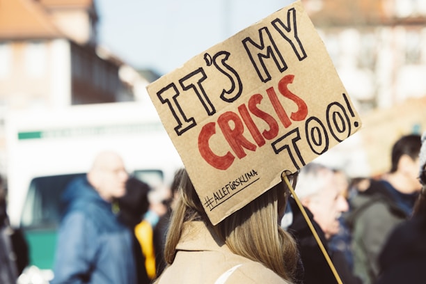 A crowd of people appears to be gathered, with a prominent sign in the foreground reading 'IT'S MY CRISIS TOO!' in bold letters. The sign is held by someone with long hair, and the text on the sign conveys a message of personal involvement or activism. The background shows more people slightly out of focus, indicating a group setting or protest.